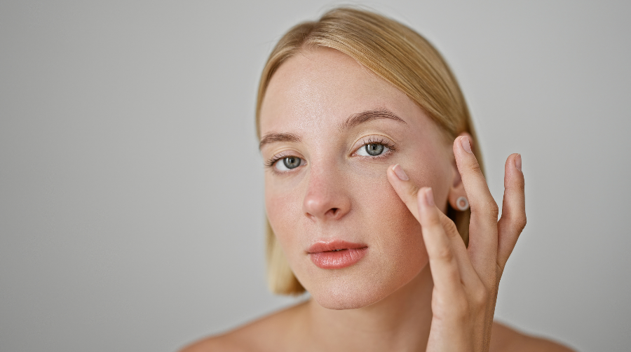 Blonde Frau trägt Creme unter das Auge auf, mit neutralem Hintergrund.
