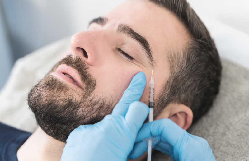 Mann mit Bart erhält Injektion im Gesicht durch blaugekleidete Hände, Nahaufnahme während kosmetischer Behandlung.